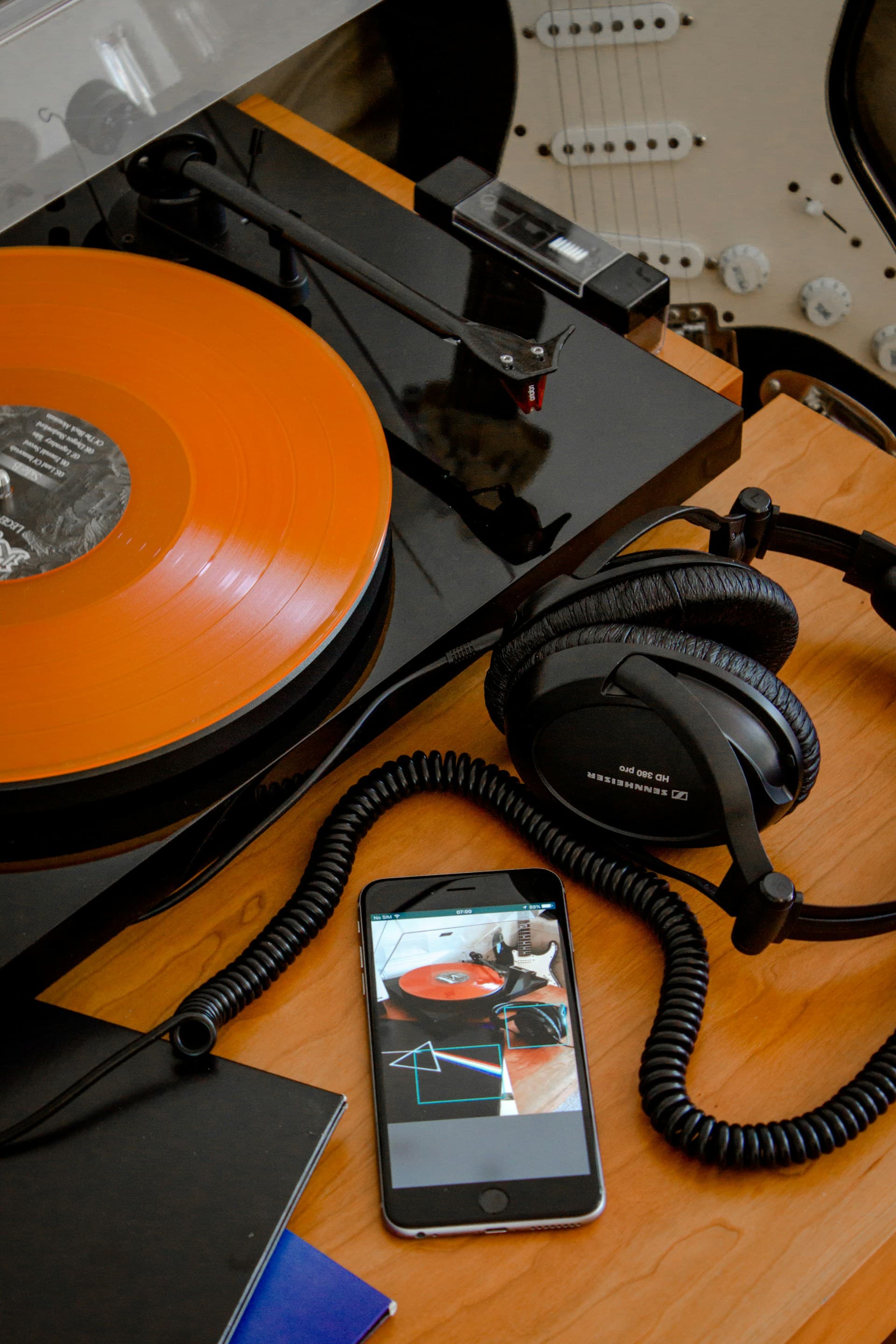 A vinyl record player sits on a table next to an iPhone, headphones, and a stack of records